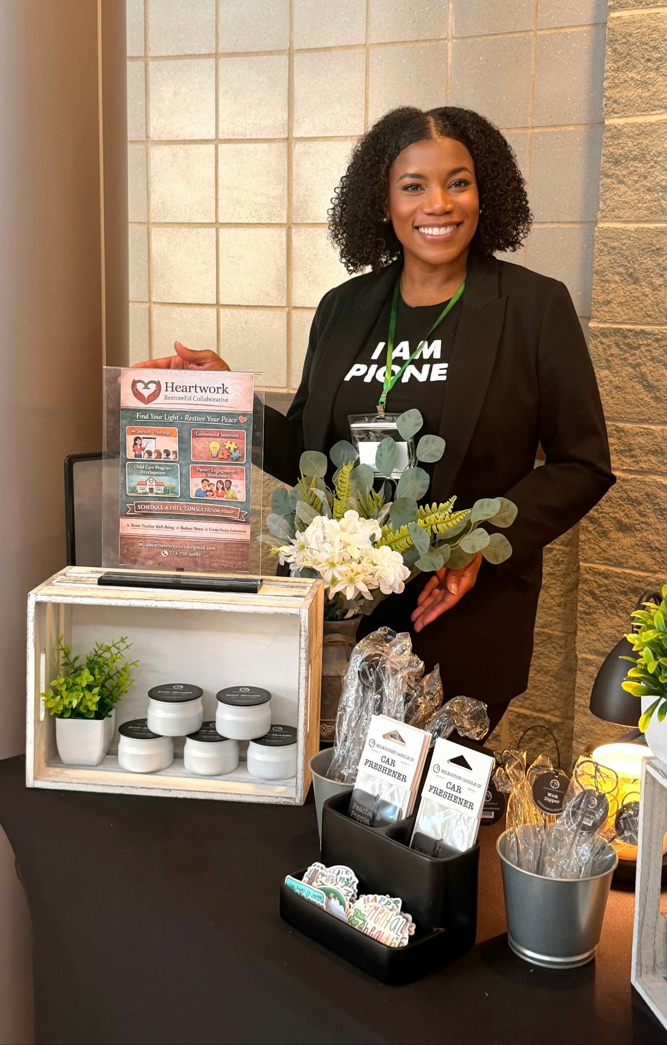 Smiling woman stands behind a table with Heartwork RestoreEd Collaborative signage and various wellness products.