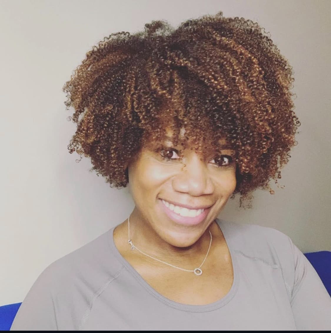Smiling woman with voluminous, highlighted curly hair wearing a grey shirt and silver necklace.